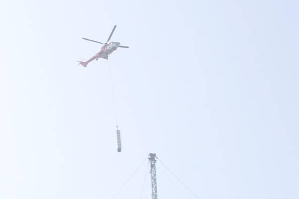 A helicopter can be seen transporting sections of the mast to the ground as workers use wires and ladders to access the upper levels of the temporary mast.