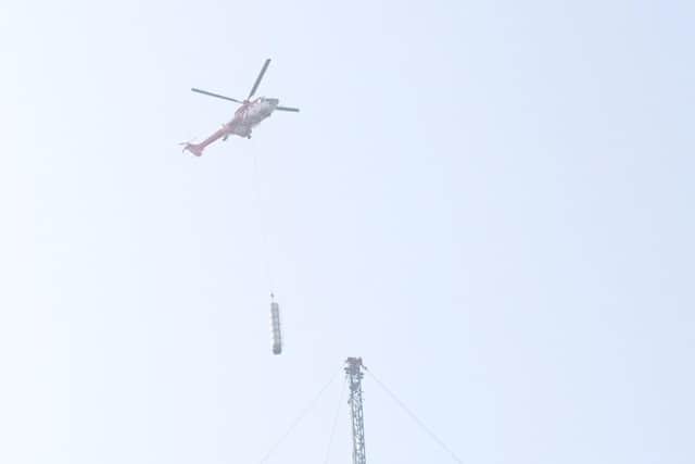 A helicopter can be seen transporting sections of the mast to the ground as workers use wires and ladders to access the upper levels of the temporary mast.